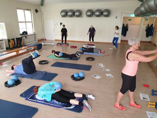 Gruppe von Personen in einem Fitnessraum, die verschiedene Übungen mit Entspannungsbällen und Matten ausführen.