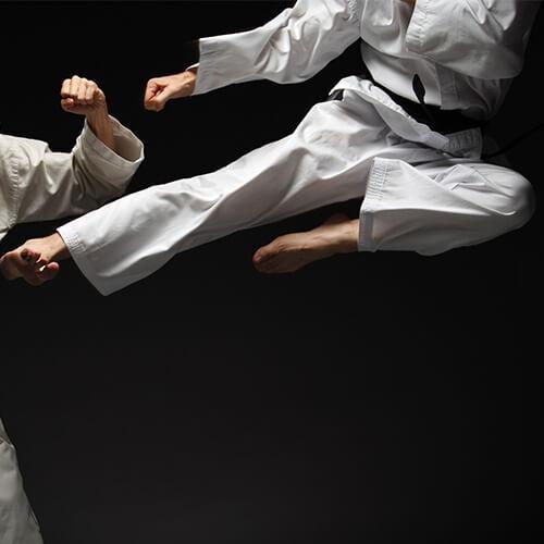 Zwei Kampfsportler in weißen Anzügen führen einen kickenden Angriff in einem dunklen Raum aus.