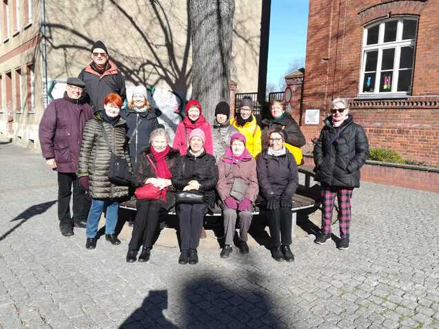 Gruppe von zwölf Personen sitzt auf einer Bank vor einem Backsteingebäude an einem sonnigen Tag.