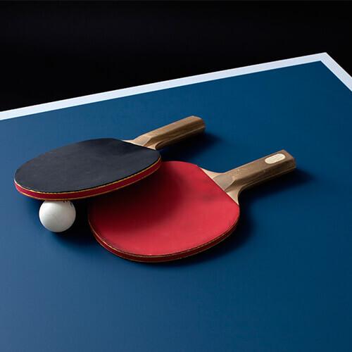 Zwei Tischtennisschläger, rot und schwarz, liegen auf einer blauen Tischtennisplatte neben einem weißen Ball.