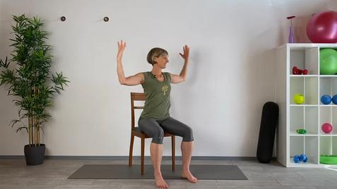 Frau sitzt auf einem Stuhl, hebt beide Arme und dehnt sich in einem hellen Raum mit Fitnessgeräten.