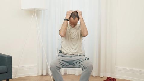 Mann sitzt auf einem Stuhl, greift mit beiden Händen an seinen Kopf und ist von einem neutralen Raum umgeben.