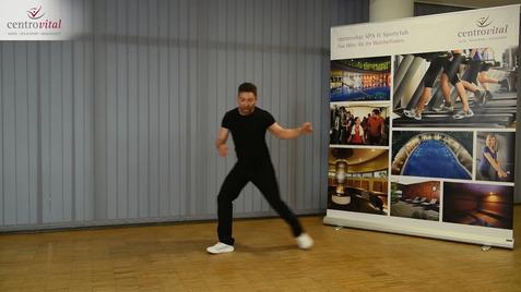 Tanzender Mann in schwarzem Outfit auf Parkettboden, mit Werbebanner im Hintergrund.