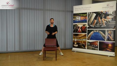 Ein Mann in sportlicher Kleidung steht hinter einem Stuhl und demonstriert eine Übung in einem Fitnessraum.