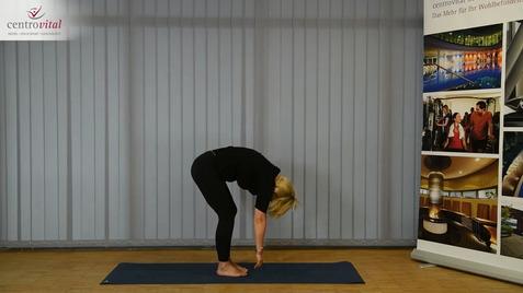 Frau in Yogahaltung mit leicht gebeugtem Oberkörper auf einer blauen Matte in einem Raum mit vertikalen Jalousien.