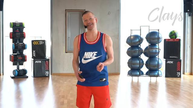 Mann in Sportbekleidung mit Mikrofon steht in einem modernen Fitnessraum mit Trainingsgeräten im Hintergrund.