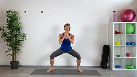 Frau in Sportkleidung steht im Squat auf einer Yoga-Matte vor einer Wand mit Pflanzen und Trainingsgeräten.