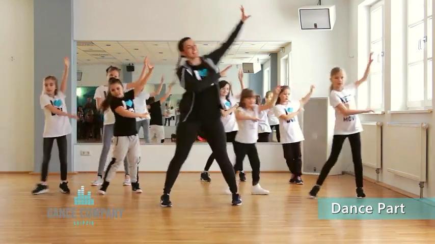 Eine Tanzgruppe mit Kindern in weißen T-Shirts übt synchron in einem Raum mit Spiegeln und Holzfußboden.