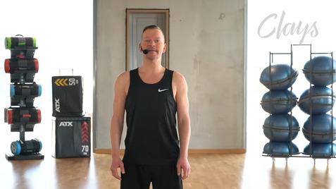 Mann in Sportkleidung mit Mikrofon steht in einem Fitnessraum vor Trainingsgeräten und einer Wand.