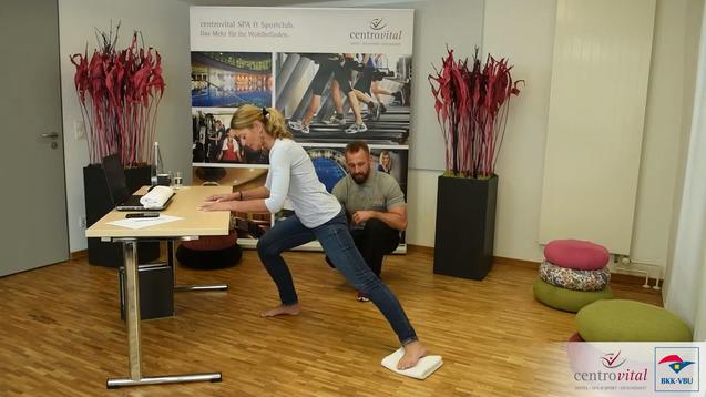 Frau führt eine Dehnübung an einem Tisch aus, während ein Mann sie dabei unterstützt. Pflanzen und Dekoration im Hintergrund.