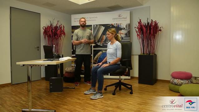 Mann im grauen T-Shirt erklärt etwas einer Frau in blauer Oberbekleidung, die auf einem Stuhl sitzt, in einem hellen Raum.