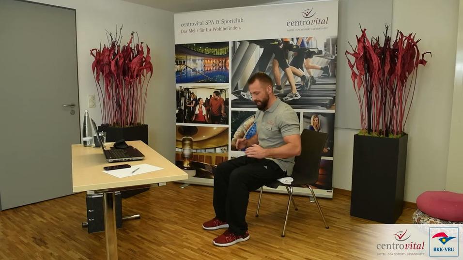 Mann sitzt auf einem Stuhl vor einem Tisch mit Laptop und Notizen, umgeben von Pflanzen und einem Werbebanner.