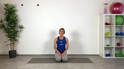 Frau in grauen Sporthosen und blauer Tanktop sitzt auf einer Yoga-Matte vor einem weißen Wandregal mit Fitnessgeräten.