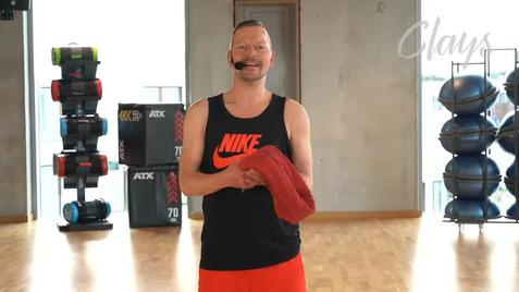 Mann mit Mikrofon in Sportkleidung hält eine runde rote Matte in einem Fitnessstudio mit Geräten im Hintergrund.
