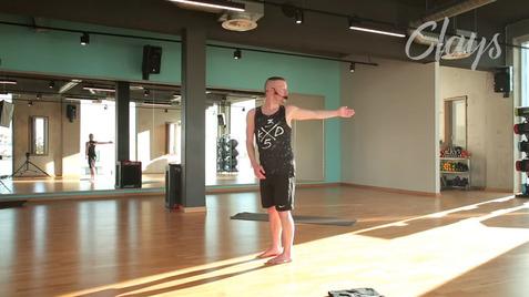 Ein Mann in Sportkleidung zeigt eine Übung in einemSportraum mit großen Fenstern und Spiegeln.