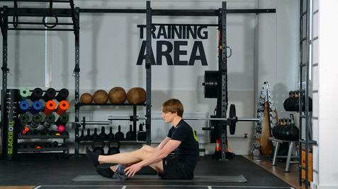 Person sitzt auf einer Matte im Fitnessstudio und dehnt die Beine; im Hintergrund Gewichte und Trainingsgeräte sichtbar.