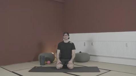 Mann im Sportoutfit sitzt auf einer Yogamatte in einem schlicht eingerichteten Raum mit roter Wand.