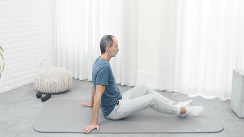 Mann sitzt auf einer Trainingsmatte, stützt sich mit den Händen ab, in einem hellen Raum mit Vorhängen.