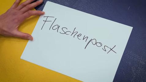 Hand, die ein Blatt Papier mit der handschriftlichen Aufschrift „Flaschenpost“ auf einem bunten Hintergrund hält.