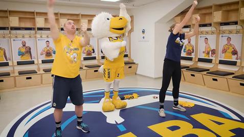 Zwei Personen und ein Maskottchen machen in einem Sportumkleideraum gemeinsame Übungen auf einem runden Logo-Boden.