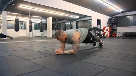 Mann in Sportkleidung führt eine Plank-Übung auf einer schwarzen Trainingsmatte in einem Fitnessstudio aus.