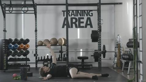 Person liegt seitlich auf dem Boden einer Trainingsfläche und streckt einen Arm zur Decke, umgeben von Fitnessgeräten.