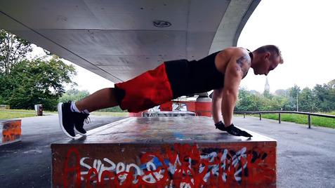 Mann führt eine Liegestütze auf einer graffiti-bedeckten Betonplatte unter einer Brücke aus.