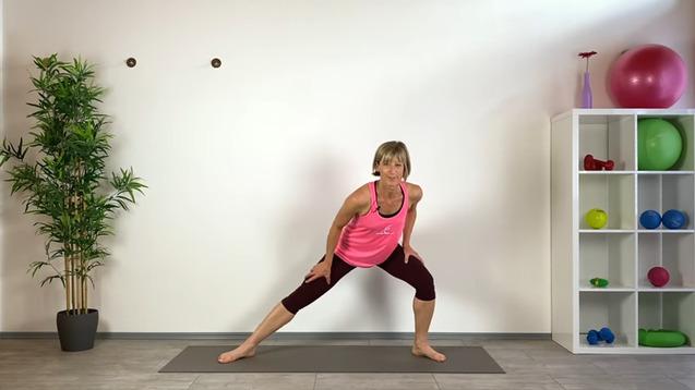 Frau in Sportkleidung macht eine Dehn- oder Fitnessübung auf einer Matte vor einer Wand mit Fitnessgeräten.