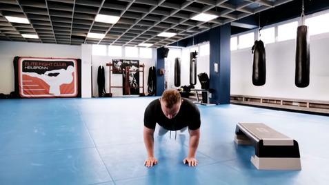 Mann macht Liegestütze auf blauem Boden in einem Fitnessstudio mit Trainingsgeräten im Hintergrund.
