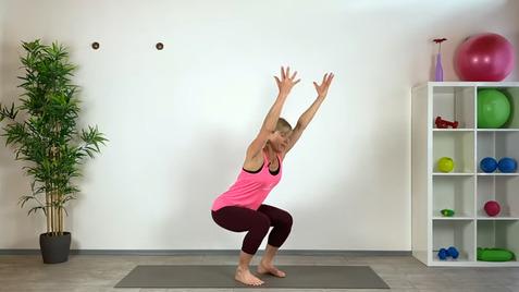 Frau in Sportkleidung macht eine Kniebeuge mit erhobenen Armen auf einer Yoga-Matte in einem hellen Raum.
