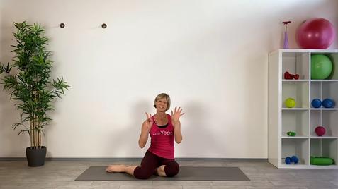 Frau im Yoga-Outfit sitzt auf einer Matte, zeigt mit beiden Händen eine Zahl, umgeben von Pflanzen und Fitnessgeräten.