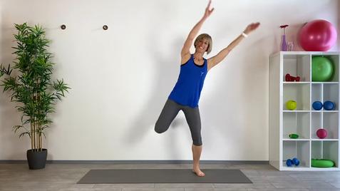 Frau in blauer Sporttop und grauen Leggings tanzt auf einer Matte, mit einem Bein angehoben. Im Hintergrund Fitnessgeräte.