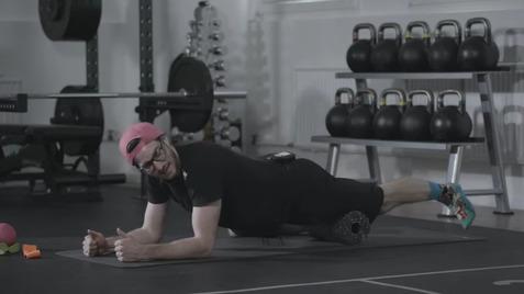 Mann in Sportkleidung liegt auf einer Matte und nutzt einen Schaumstoffroller für das Training in einem Fitnessstudio.