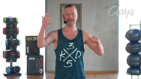 Mann in sportlichem Shirt mit Mikrofon spricht trainierend, Fitnessgeräte im Hintergrund auf einem hellen Boden.