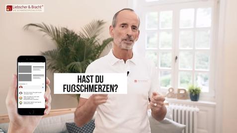 Mann mit grauem Haar trägt ein Polohemd und spricht über Fußschmerzen, während ein Smartphone in einer Hand gehalten wird.