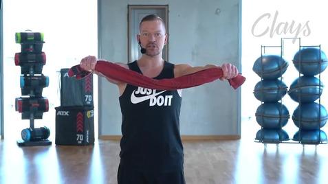 Mann demonstriert eine Übung mit einem roten Widerstandsband in einem Fitnessstudio. Sportgeräte sind im Hintergrund sichtbar.