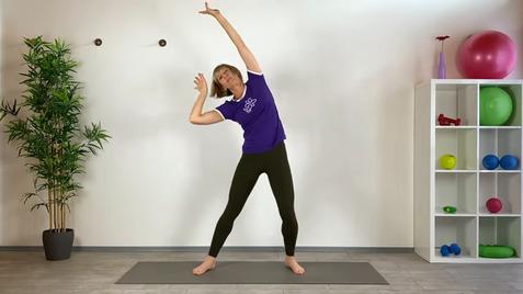 Frau in Sportkleidung dehnt sich seitlich auf einer Yogamatte vor einer weißen Wand mit Pflanzen und Fitnessgeräten.