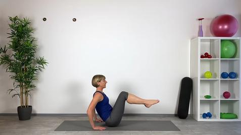 Frau in Yogaposition auf Matte, mit angehobenen Beinen, in einem Raum mit Fitnessgeräten und Pflanzen im Hintergrund.