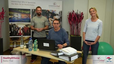 Drei Personen stehen und sitzen vor einem Tisch mit Laptop und Wasserflaschen, umgeben von Pflanzen und Sportgeräten.