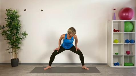 Frau in Sportkleidung macht eine Übung auf einer Matte in einem hellen Raum mit Fitnessgeräten im Hintergrund.