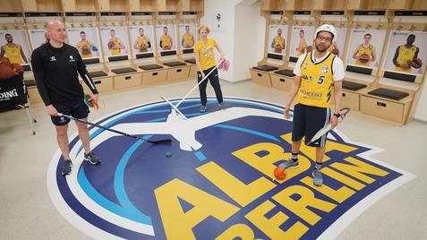 Drei Personen stehen auf einem ALBA Berlin-Logo im Umkleideraum, ausgestattet mit Sportgeräten wie Schlägern und Bällen.