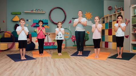 Sechs Personen in Yogastellungen auf Matten im Innenraum, umgeben von bunten Möbeln und Spielzeug.