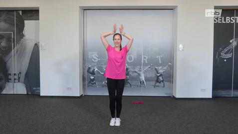 Frau in pinkem T-Shirt steht mit erhobenen Armen vor einer Wand mit Sportmotiven.