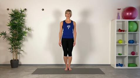 Frau steht barfuß auf einer Yogamatte vor einer weißen Wand, umgeben von Pflanzen und Trainingsgeräten.