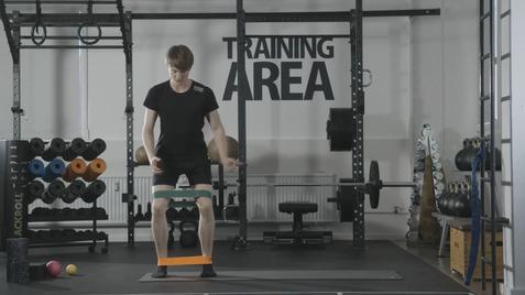 Junger Mann trainiert in einem Fitnessbereich mit Widerstandsbändern an den Oberschenkeln. Ausstattung im Hintergrund sichtbar.