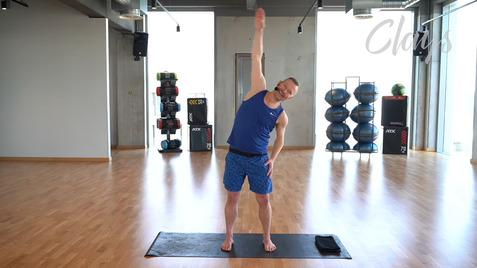 Mann in Sportkleidung, der beim Dehnen auf einer Matte steht, mit einem Arm hochgestreckt in einem Fitnessstudio.