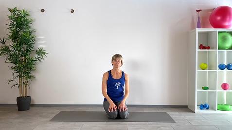 Eine Frau in Sportkleidung kniet auf einer Matte in einem hellen Raum mit Pflanzen und Trainingsgeräten im Hintergrund.