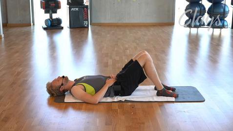 Mann liegt auf einer Matte im Fitnessraum mit Handtuch, Beine angewinkelt und Hände auf dem Bauch.