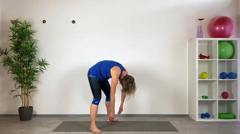 Frau in blauer Sportbekleidung beugt sich über eine Yogamatte, um ihre Zehen zu erreichen; Fitnessgeräte im Hintergrund.