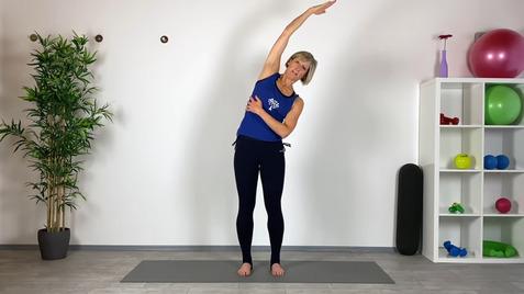 Woman in blue tank top performs a side stretch on a yoga mat in a fitness studio with exercise equipment in the background.